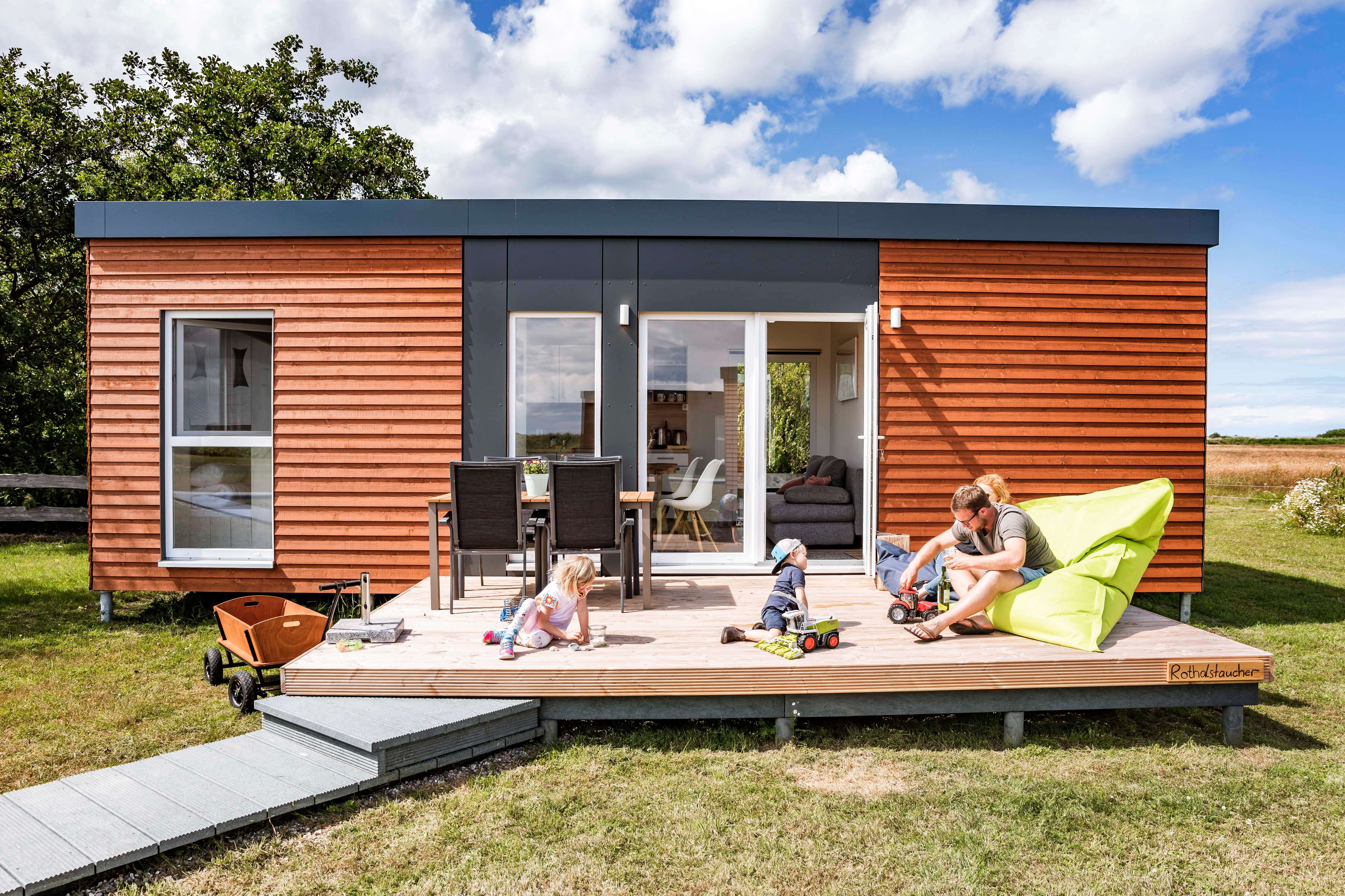 Junge Familie sitzt auf der Terrasse vor einem modernen Tinyhouse auf einem Campingplatz