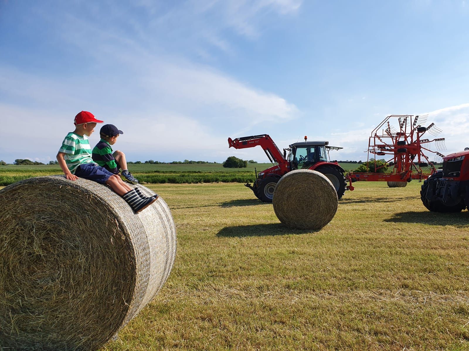 Kinder sitzen auf einem Heuballen auf einem Feld und beobachten einen Trecker bei der Heuernte