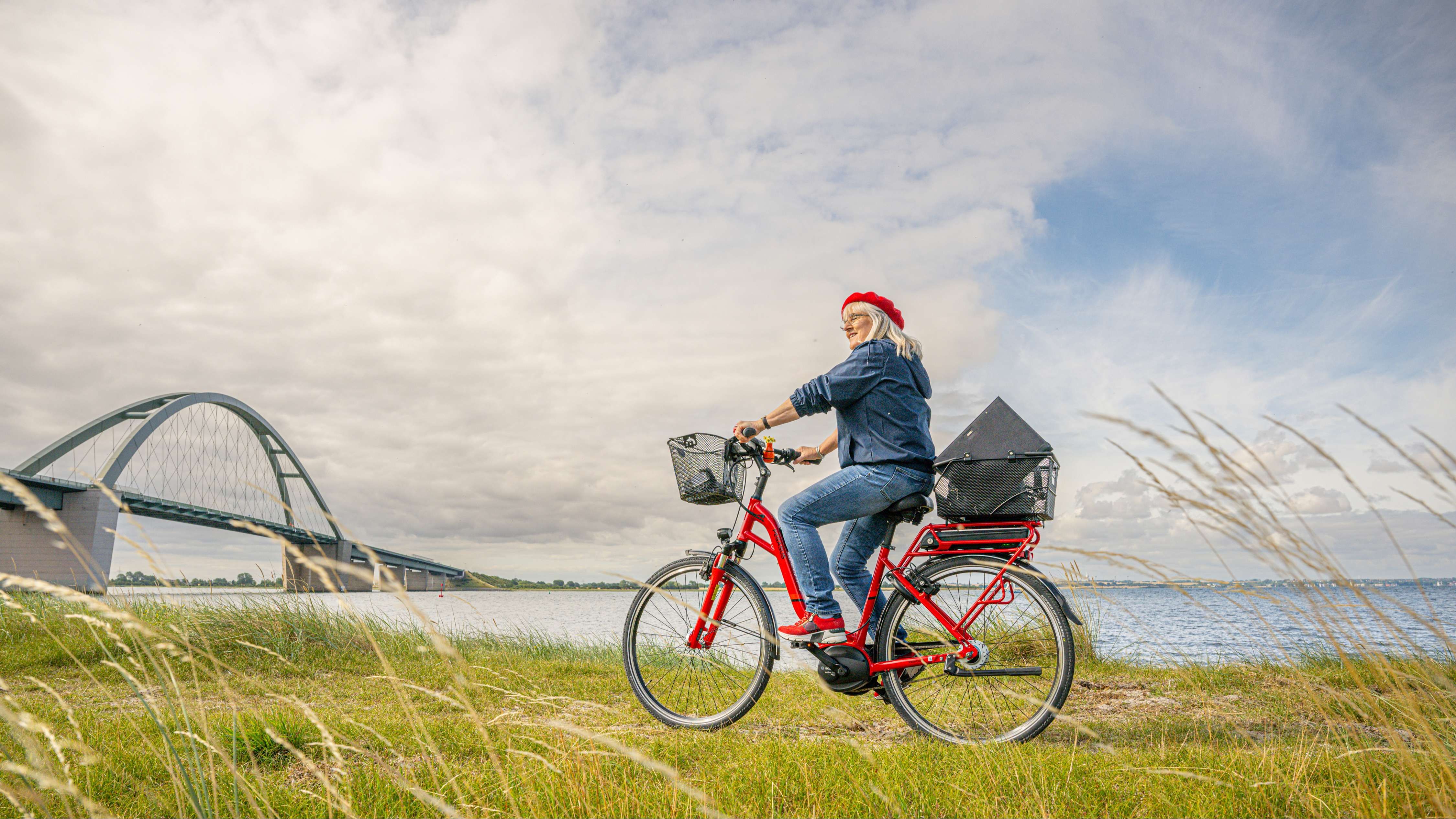 Eine Frau fährt mit ihrem roten Fahrrad auf einer Wiese nahe der Ostsee entlang, im Hintergrund sieht man die Fehmarnsundbrücke.